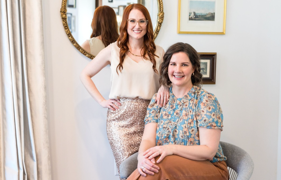 Two women posing together in a room with a mirror and framed pictures on the wall.